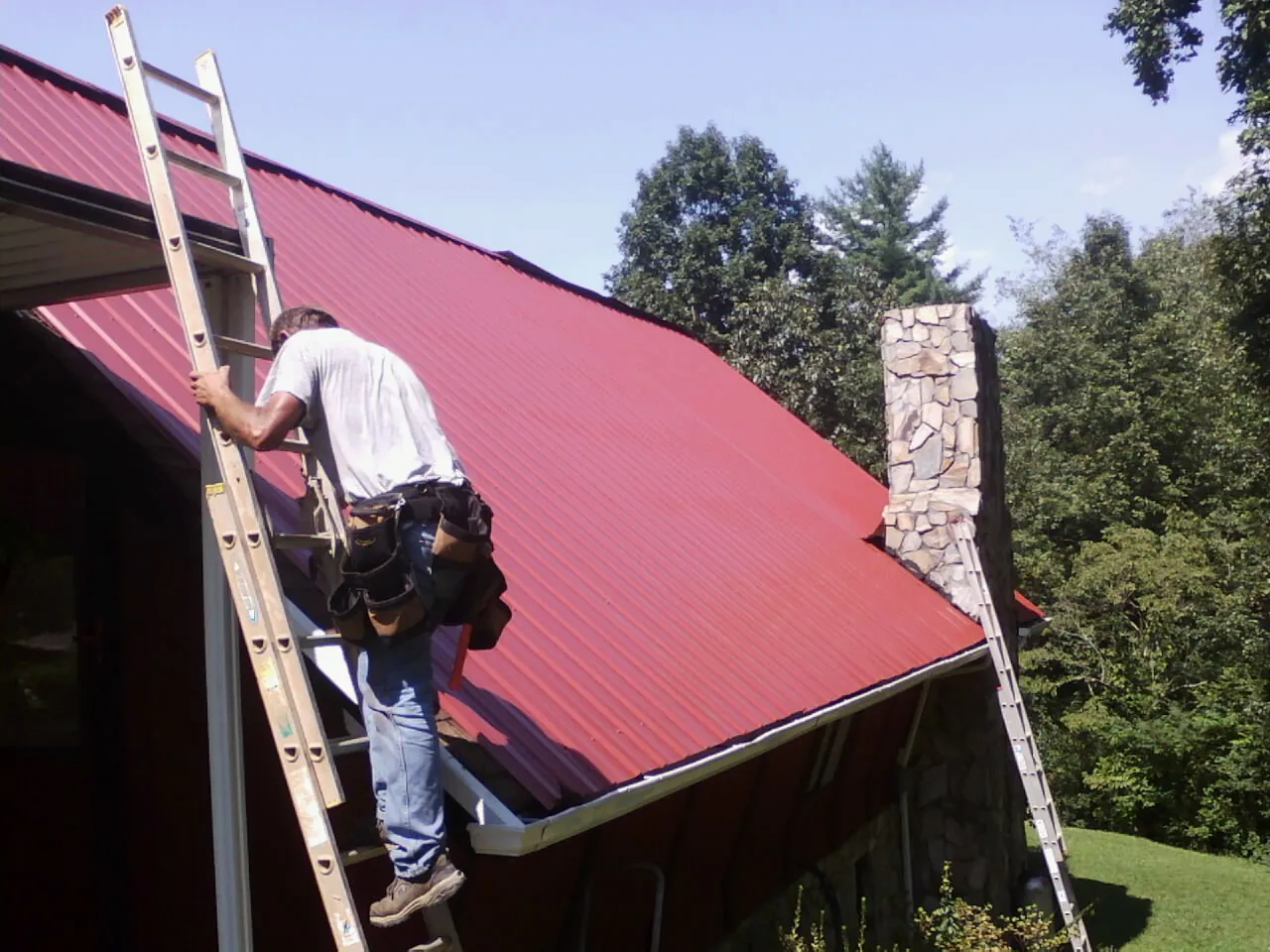 Roofer on a ladder beside a red metal roof and stone chimney — Soffit & Fascia Repair by Roofing Brothers in Hodges